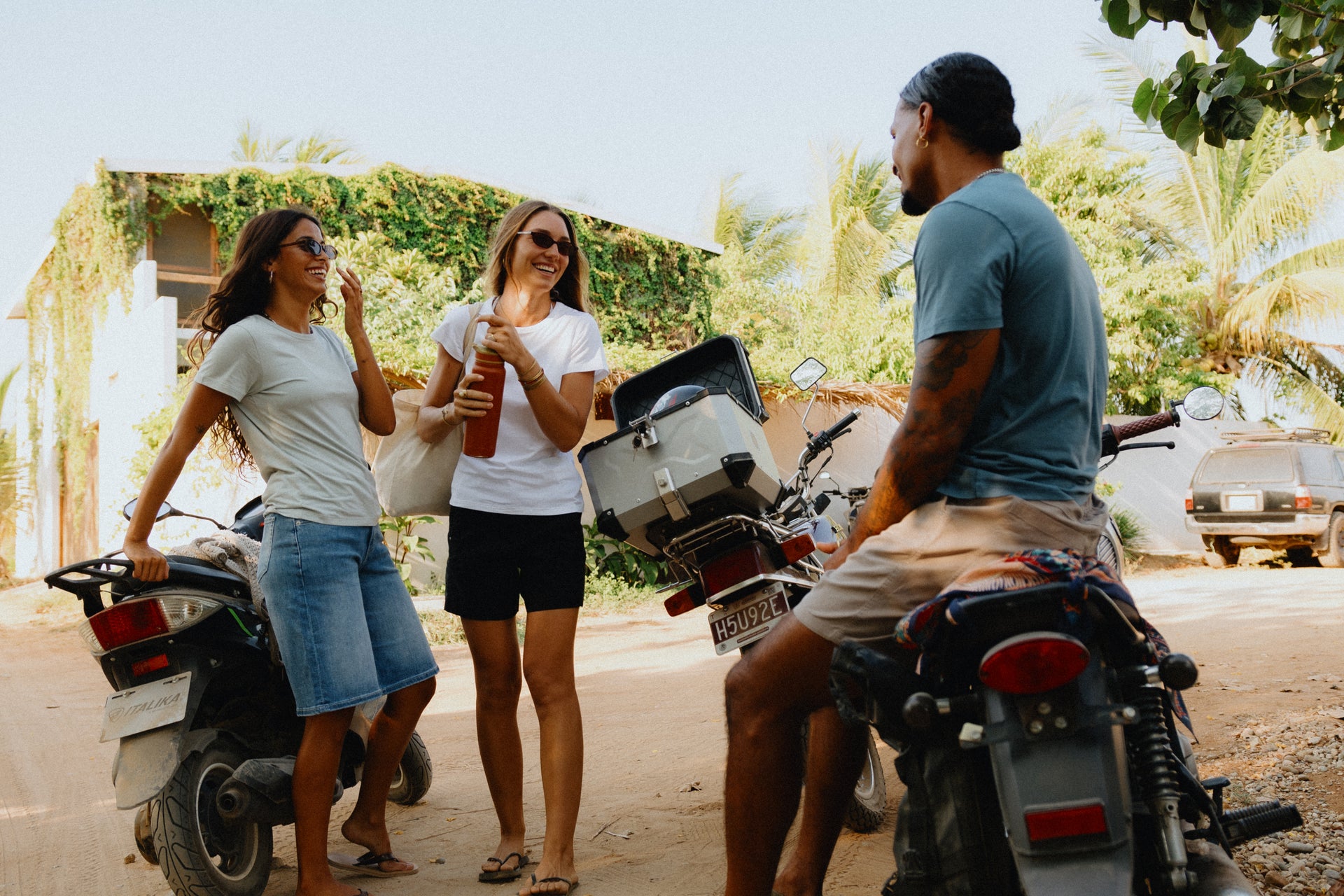 Three people standing outdoors between two motorcycles in a sunny, tropical setting. One man is seated on a motorcycle on the right, and two women stand on the left, one holding a drink. They all wear DUER hot weather clothing such as DUER no sweat shorts and Live Lite denim shorts