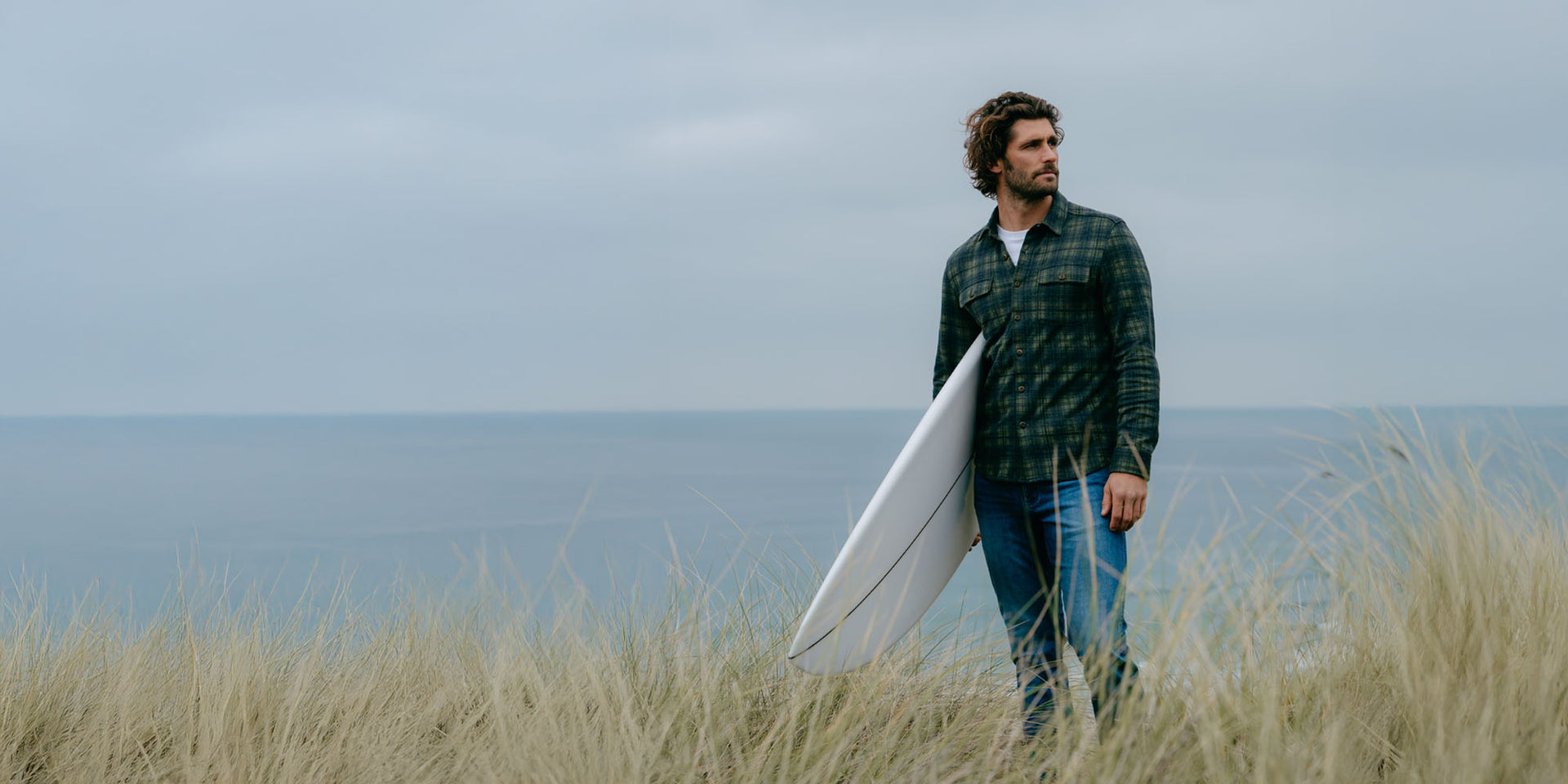 Man holding a surfboard standing in tall grass near the ocean wearing DUER mens jeans