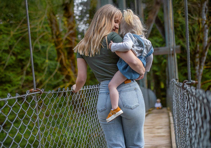 Woman holding a child on a wooden suspension bridge with greenery in the background wearign Duer Womens jeans