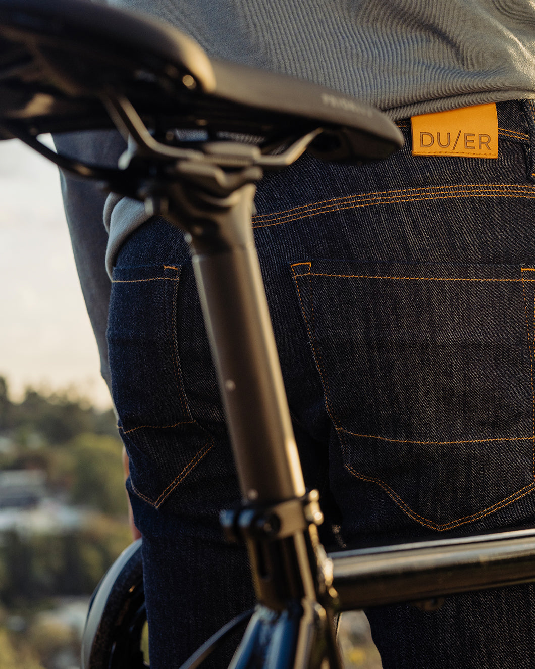 Close-up of the back of a person in dark denim jeans with a yellow DU/ER tag on the waistband, standing next to a bike.