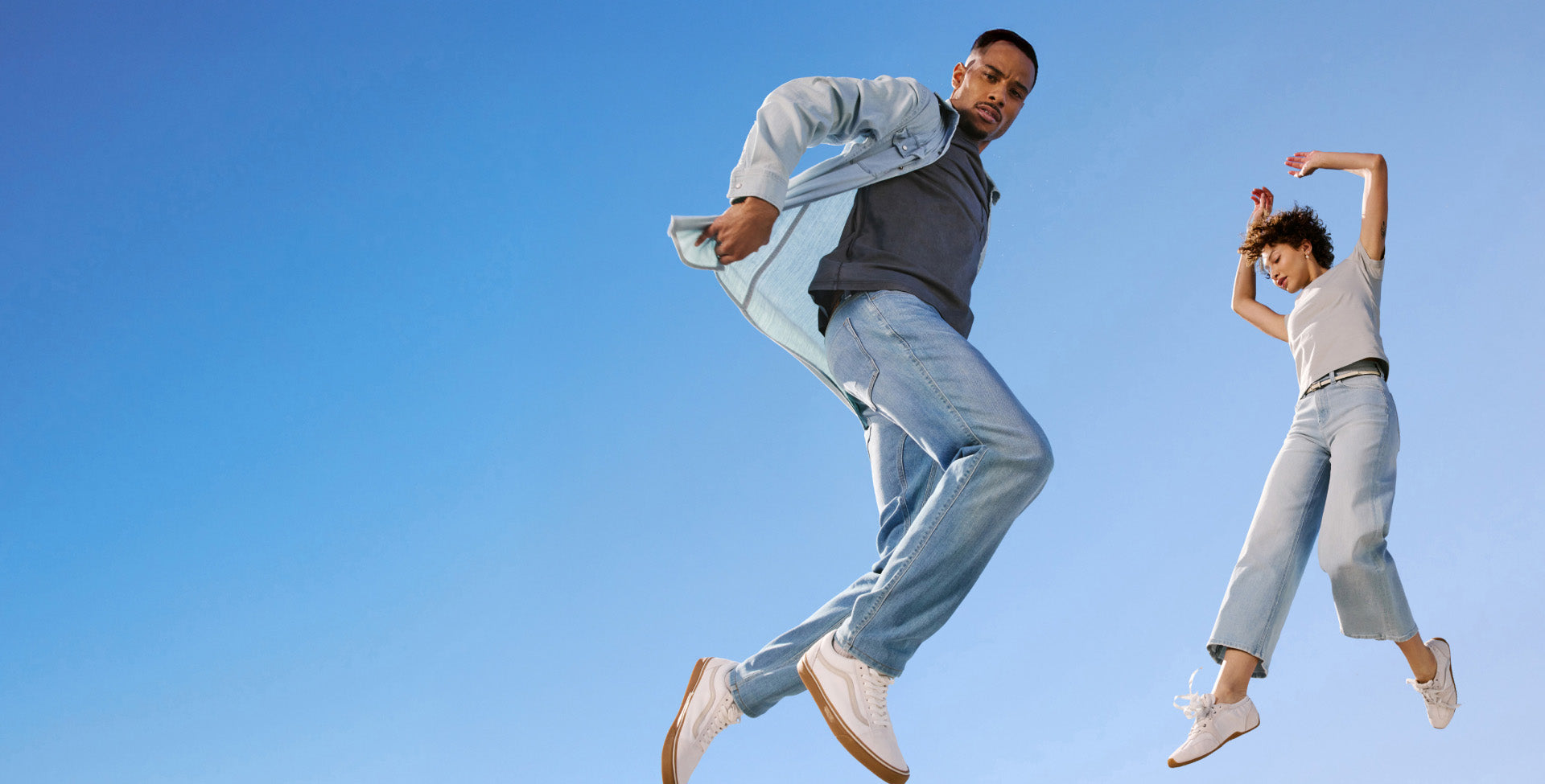 Two people jumping against a clear blue sky wearing DUER Denim and clothing