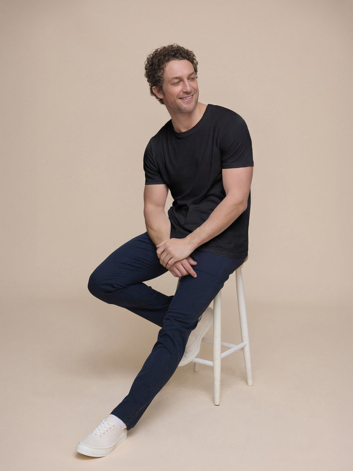 Man sitting on a stool wearing a black t-shirt and DUER navy chino pants against a beige background