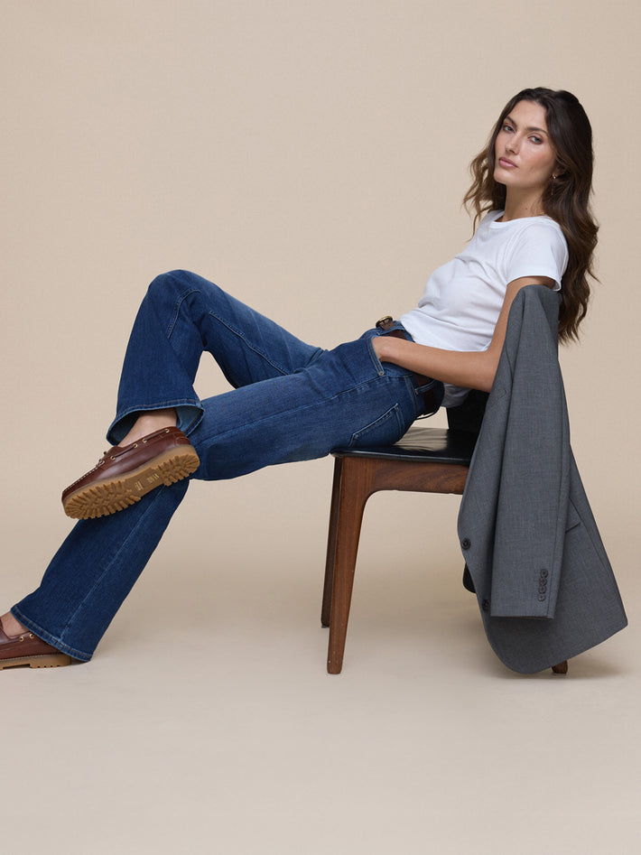 Woman sitting on a chair wearing blue DUER performance jeans and a white shirt, with a gray jacket draped over the backrest.